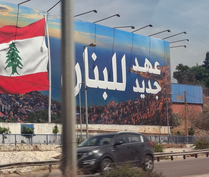 A car drives in front of a poster which displays an aerial image of the Bay of Jounieh, with text and an image of the Lebanese flag superimposed on it. The text reads: عهد جديد للبنان [translates to: A New Era for Lebanon]