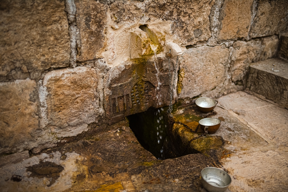 Water falls from a spout in the brick wall into a hole in the ground. Three silver bowls lie around it.