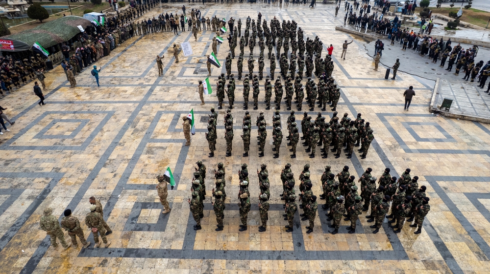 Soldiers stand in six orderly groups behind soldiers holding the Syria flag